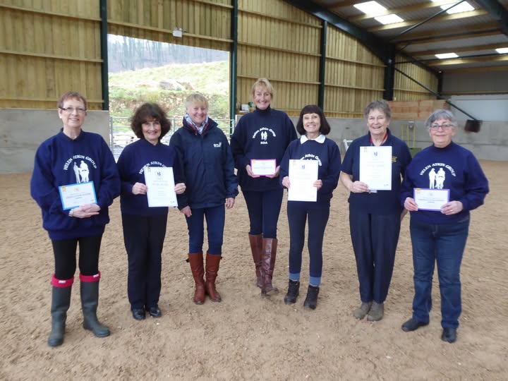 A group of smiling volunteers stand in a covered arena holding their long service award certificates and medals. They are dressed smartly, with several people in the centre and others around them, celebrating their recognition for years of service. The wider setting is a horse-riding arena with a bright, welcoming atmosphere. Text in the image reads Long service awards 2022, and the overall mood is proud and joyful.